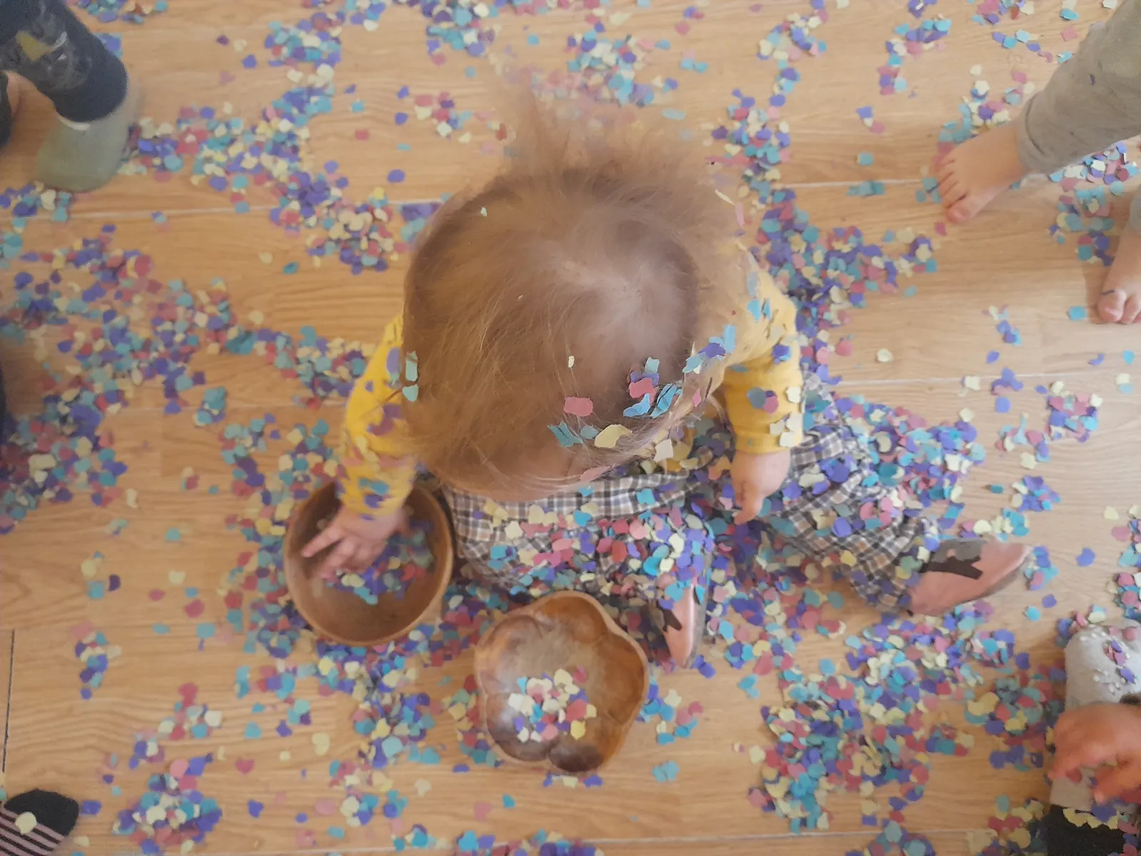 Un infant assegut al terra envoltat de confeti de colors, amb bols de fusta