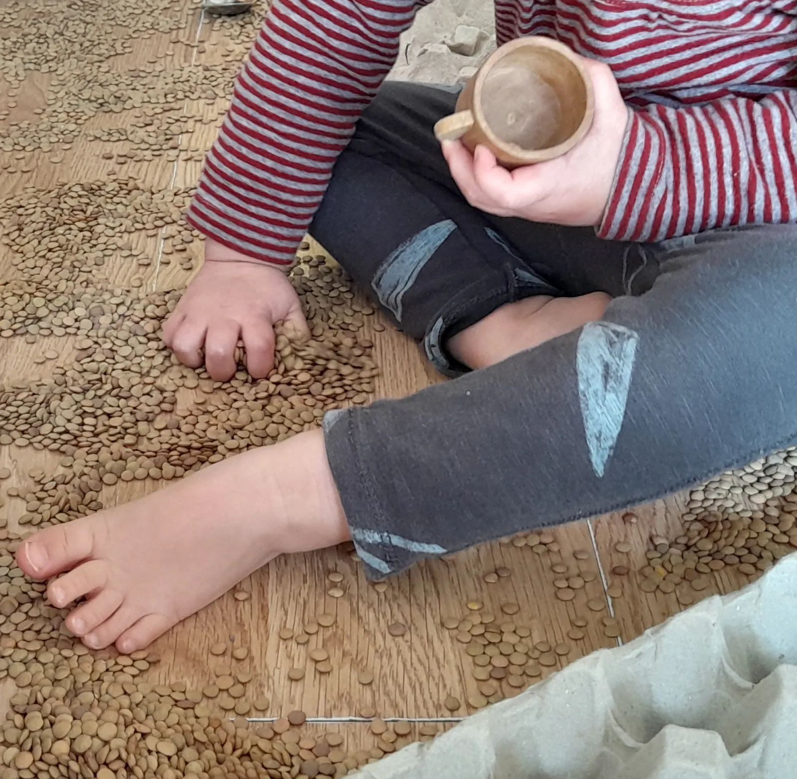 Un infant descalç assegut sobre llenties escampades, amb una tassa de fusta a la mà