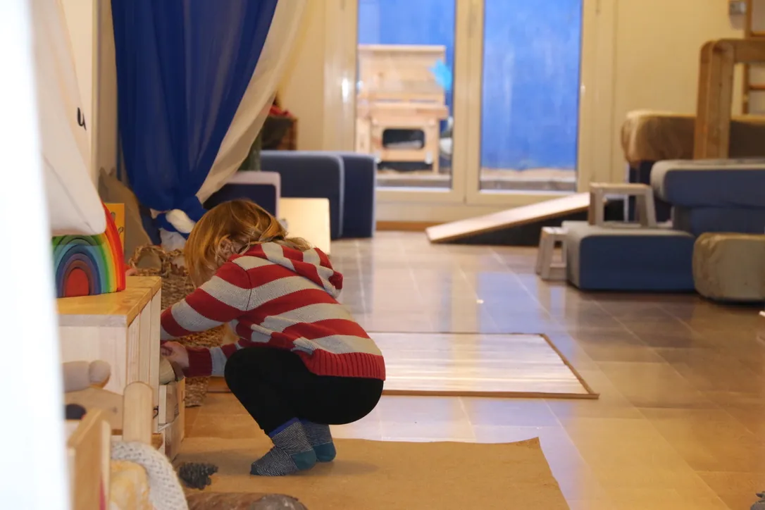 Una acompanyant ajupida ordenant materials de joc al fons d’una sala amb finestral